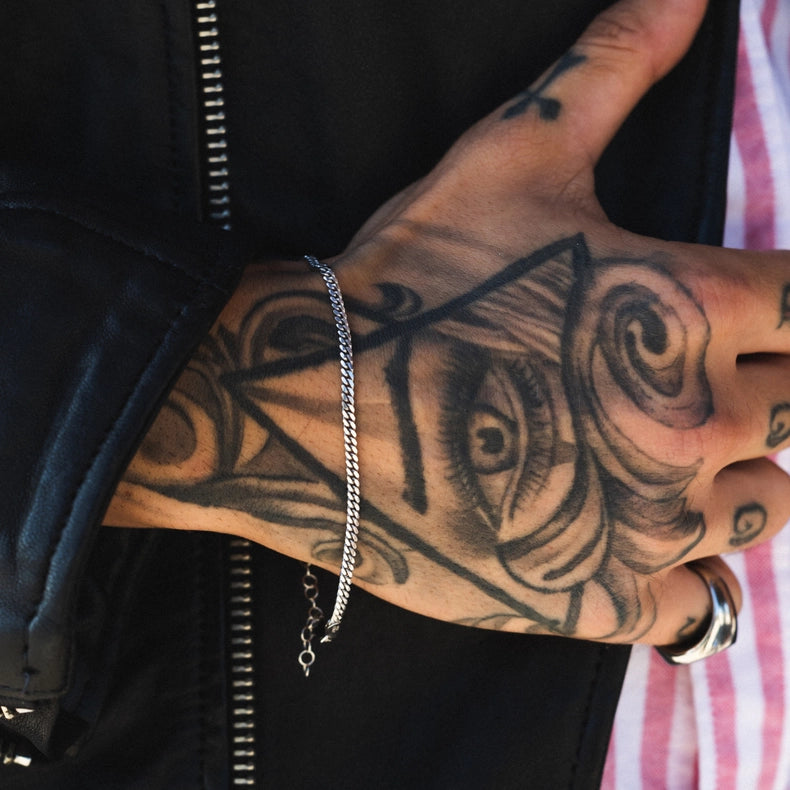 Hand with tattoos wearing a 925 sterling silver Cuban chain bracelet and ring, against a black jacket background.