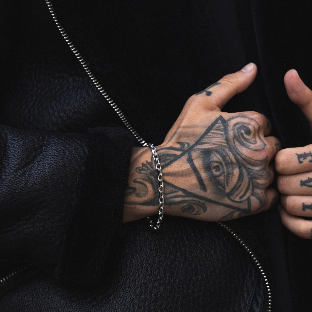 A man with tattoos wearing a 925 sterling silver rolo chain bracelet against a dark jacket