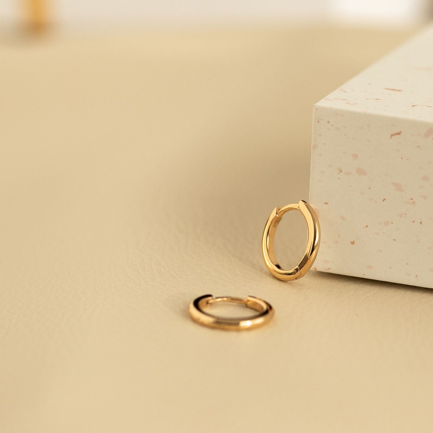 Two 18k gold-plated hoop earrings on a beige surface with a white box in the background