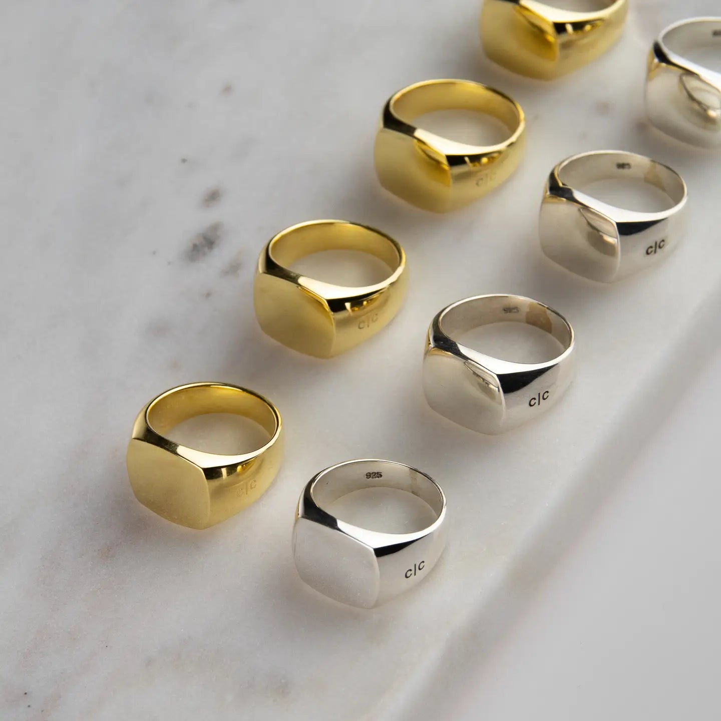 Row of men's 925 sterling silver signet rings next to 18k gold plated signet rings on a white marble worktop